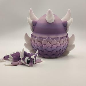 May include: A purple and white dragon egg with a matching baby dragon figurine. The egg has a layered design with white wings and horns. The baby dragon is lying down, also in purple and white.