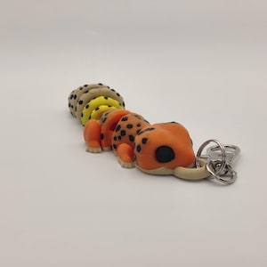 May include: A colorful caterpillar keychain. The keychain features an orange body with black spots, yellow and tan segments, and a cream-colored head. The keychain is attached to a silver keyring.