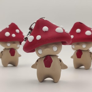May include: Three mushroom-shaped figurines with red caps dotted with white spots. The figurines have tan bodies, small arms, and a red diamond-shaped detail on their chests. Each figure has a keychain attached to the top of the mushroom cap.