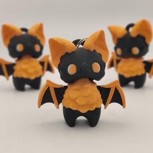 May include: Three small bat figurines. Each bat has a black body, orange wings and ears, and orange chest. The eyes are white. The bats are standing on a white surface. The bats are keychains.