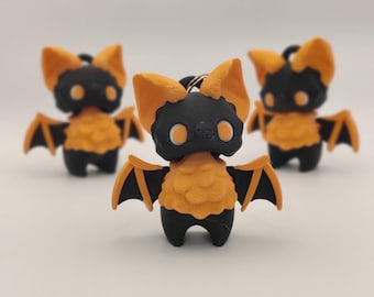 May include: Three small bat figurines. Each bat has a black body, orange wings and ears, and orange chest. The eyes are white. The bats are standing on a white surface. The bats are keychains.