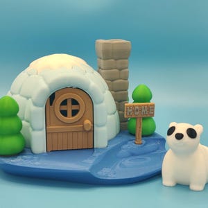 May include: A whimsical 3D-printed diorama featuring an igloo with a brown door, a chimney, and a sign that reads "HOME." A white polar bear with black eyes sits nearby. Green trees and a blue base complete the scene.