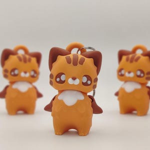 May include: Orange and brown cat-shaped keychain figurines with white accents. The cat figurines have large eyes and are standing upright. The keychains have a ring at the top for attaching to keys or bags. The background is a soft, neutral color.