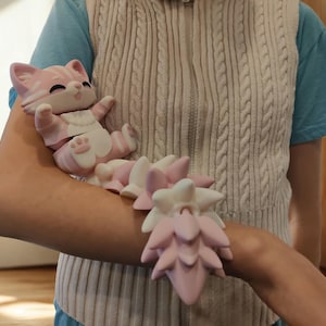 May include: A pink and white cat figurine with a matching tail accessory. The cat is lying down with its arms outstretched. The tail is a series of layered, petal-like shapes. The cat is wearing a white bib.