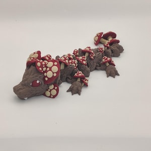 May include: A whimsical, brown dragon figurine adorned with red and white spotted mushroom accents. The creature has a detailed face with a single eye and is posed on a white surface. The overall design is fantasy-themed.