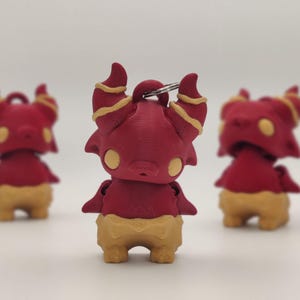 May include: Three red and tan figurines of a cartoonish creature with horns, wings, and a round body. The figurines have yellow accents on the horns and belly. The figurines are standing on a white surface.