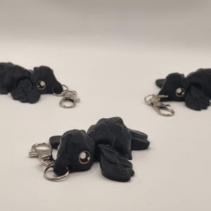 May include: Three black crow keychains with silver-colored clasps and key rings are displayed against a white background. Each crow has a detailed, textured design and a small, round, light-colored eye.