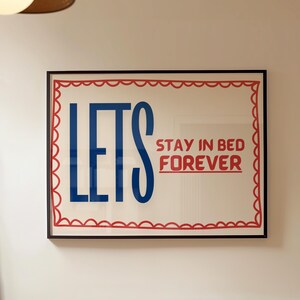 May include: A framed print with a red and white scalloped border. The print features the text "Let's Stay in Bed Forever" in blue and red lettering.