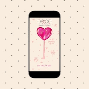 Puede incluir: La pantalla de un teléfono inteligente que muestra una piruleta rosa en forma de corazón en un palo con un fondo blanco y copos de nieve rosas. La hora es 08:00 y la fecha es domingo 15 de abril. El texto "i'm just a girl" se muestra en la parte inferior de la pantalla.