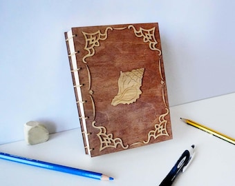 Puede incluir: Un diario rectangular de madera con acabado marrón oscuro, que presenta un emblema de concha cortado con láser en el centro y filigranas decorativas en las esquinas. El lomo está encuadernado con cordón visible de color claro.