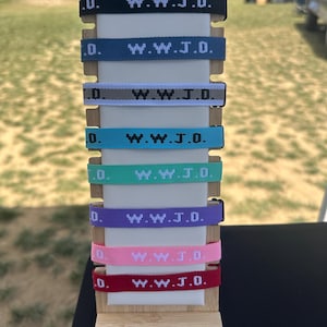 May include: A display of colorful woven bracelets with the text "W.W.J.D." in white. The bracelets are in various colors, including black, blue, gray, teal, green, purple, pink, and red. The bracelets are displayed on a wooden stand.