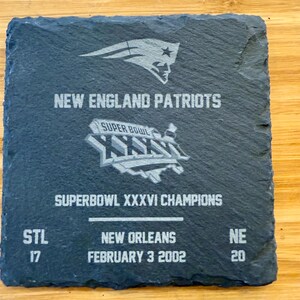 May include: A black slate coaster with a silver graphic of the New England Patriots logo, the Super Bowl XXXVI logo, and the text "NEW ENGLAND PATRIOTS", "SUPER BOWL XXXVI CHAMPIONS", "STL 17", "NEW ORLEANS", "FEBRUARY 3 2002", and "NE 20".