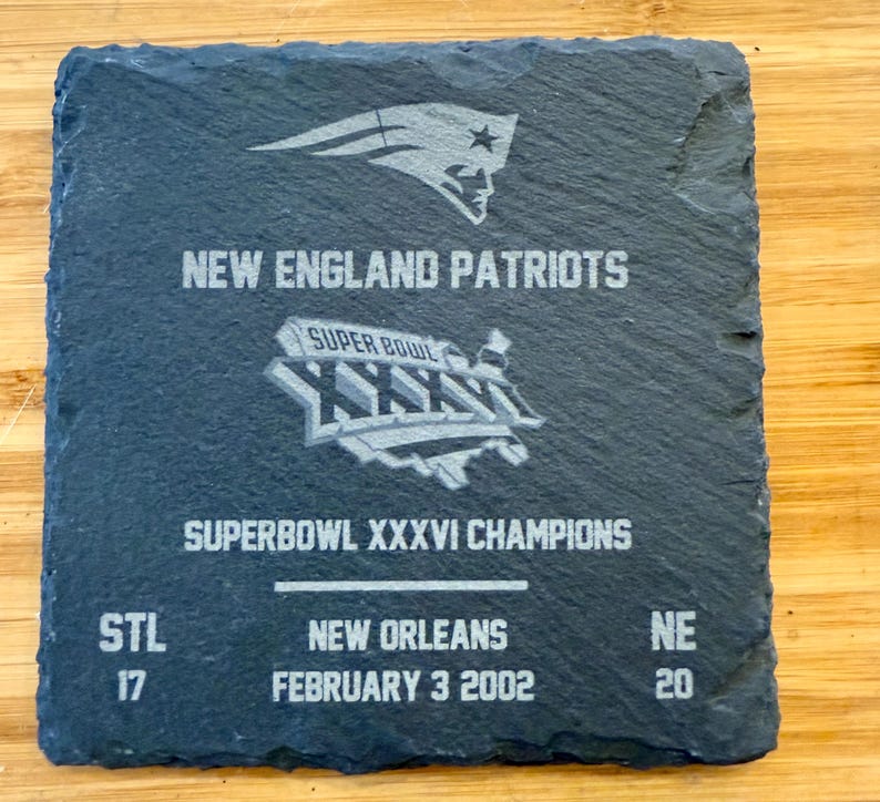 May include: A black slate coaster with a silver graphic of the New England Patriots logo, the Super Bowl XXXVI logo, and the text "NEW ENGLAND PATRIOTS", "SUPER BOWL XXXVI CHAMPIONS", "STL 17", "NEW ORLEANS", "FEBRUARY 3 2002", and "NE 20".