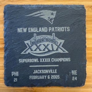 May include: A black slate coaster with a silver New England Patriots Super Bowl XXXIX logo. The text "SUPER BOWL XXXIX CHAMPIONS" is printed below the logo. The score of the game is printed below the text: "PHI 21 NE 24". The date of the game is printed below the score: "FEBRUARY 6 2005".
