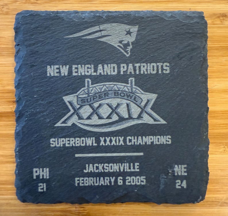 May include: A black slate coaster with a silver New England Patriots Super Bowl XXXIX logo. The text "SUPER BOWL XXXIX CHAMPIONS" is printed below the logo. The score of the game is printed below the text: "PHI 21 NE 24". The date of the game is printed below the score: "FEBRUARY 6 2005".
