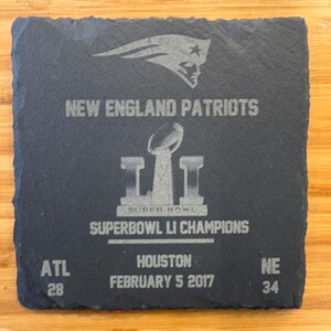 May include: A black slate coaster with a silver engraved image of the New England Patriots logo, the Super Bowl LI trophy, and the text "NEW ENGLAND PATRIOTS", "SUPER BOWL", "SUPERBOWL LI CHAMPIONS", "ATL 28", "HOUSTON FEBRUARY 5 2017", and "NE 34".