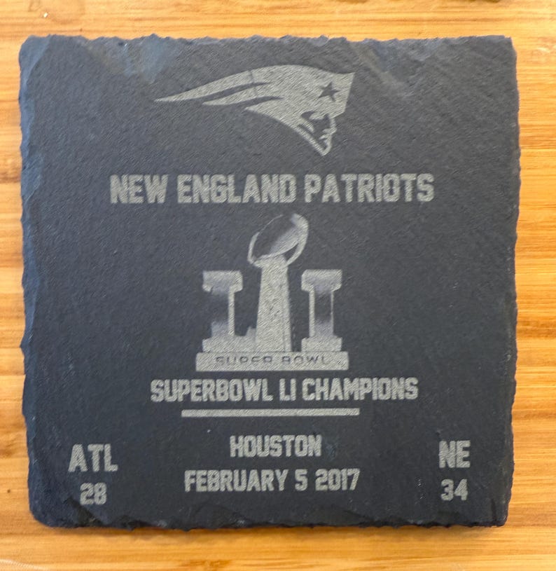 May include: A black slate coaster with a silver engraved image of the New England Patriots logo, the Super Bowl LI trophy, and the text "NEW ENGLAND PATRIOTS", "SUPER BOWL", "SUPERBOWL LI CHAMPIONS", "ATL 28", "HOUSTON FEBRUARY 5 2017", and "NE 34".