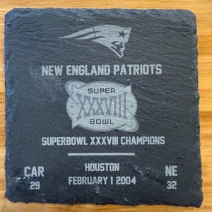 May include: A black slate coaster with a silver design commemorating the New England Patriots Super Bowl XXXVII victory. The coaster features the team logo, the Super Bowl logo, and the score of the game: CAR 29, NE 32. The text "SUPERBOWL XXXVII CHAMPIONS" and "FEBRUARY 1 2004" are also included.