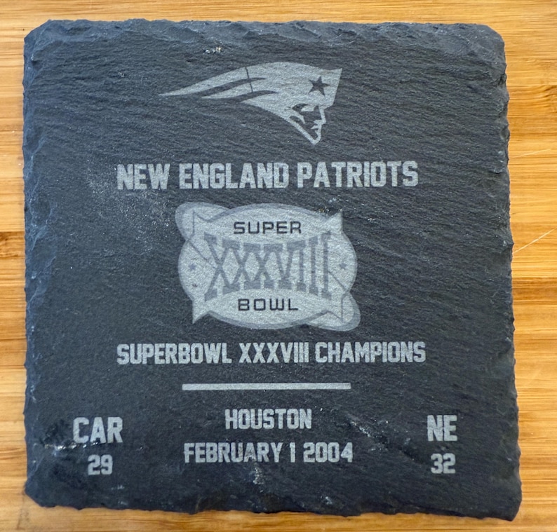May include: A black slate coaster with a silver design commemorating the New England Patriots Super Bowl XXXVII victory. The coaster features the team logo, the Super Bowl logo, and the score of the game: CAR 29, NE 32. The text "SUPERBOWL XXXVII CHAMPIONS" and "FEBRUARY 1 2004" are also included.