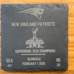 May include: A black slate plaque with the words "New England Patriots" and "Super Bowl XLIX Champions" engraved on it. The plaque also includes the date "February 1 2015" and the scores "SEA 24" and "NE 28".
