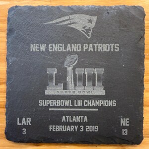 May include: A black slate coaster with a silver design commemorating the New England Patriots Super Bowl LIII victory. The coaster features the team logo, the Super Bowl LIII trophy, and the score of the game: LAR 3, NE 13. The text "Super Bowl LIII Champions" and "February 3, 2019" are also included.