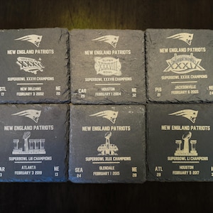 May include: Six black slate coasters with white text and images commemorating the New England Patriots Super Bowl wins. Each coaster features the team logo, the Super Bowl number, the year, and the opponent city.
