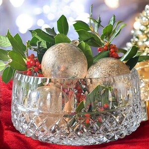 Puede incluir: Un cuenco de cristal tallado lleno de adornos navide&ntilde;os dorados, hojas verdes y bayas rojas. El cuenco se encuentra sobre una superficie roja, con un fondo borroso de luces y un peque&ntilde;o &aacute;rbol de Navidad.