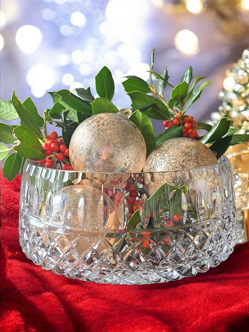 Puede incluir: Un cuenco de cristal tallado lleno de adornos navide&ntilde;os dorados, hojas verdes y bayas rojas. El cuenco se encuentra sobre una superficie roja, con un fondo borroso de luces y un peque&ntilde;o &aacute;rbol de Navidad.