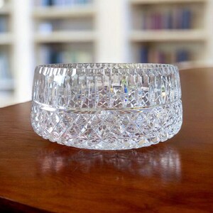Puede incluir: Un cuenco de cristal tallado transparente con un patr&oacute;n de diamantes en la mitad inferior y cortes verticales en la mitad superior. El cuenco est&aacute; sobre una superficie de madera oscura. El fondo est&aacute; borroso, mostrando una estanter&iacute;a.