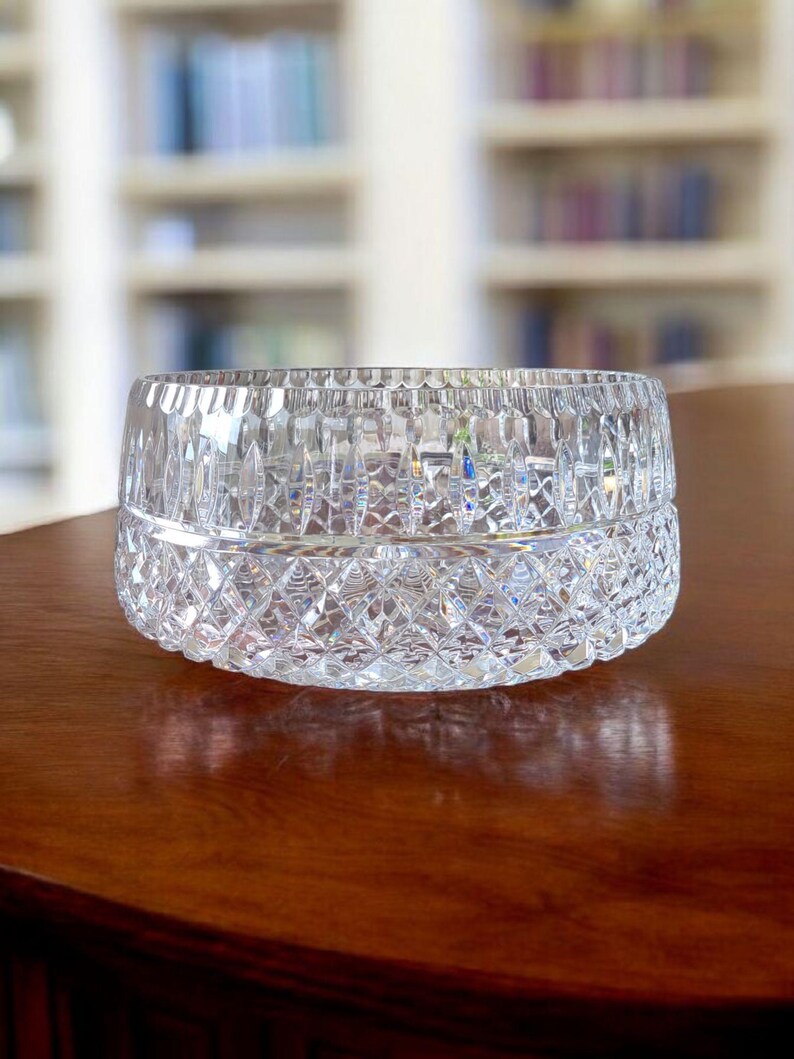 Puede incluir: Un cuenco de cristal tallado transparente con un patr&oacute;n de diamantes en la mitad inferior y cortes verticales en la mitad superior. El cuenco est&aacute; sobre una superficie de madera oscura. El fondo est&aacute; borroso, mostrando una estanter&iacute;a.