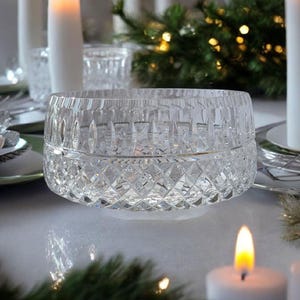 May include: A clear, cut-crystal bowl with a diamond pattern. The bowl sits on a white surface, surrounded by lit white candles and evergreen sprigs, suggesting a festive or holiday theme. The bowl's intricate design catches the light.