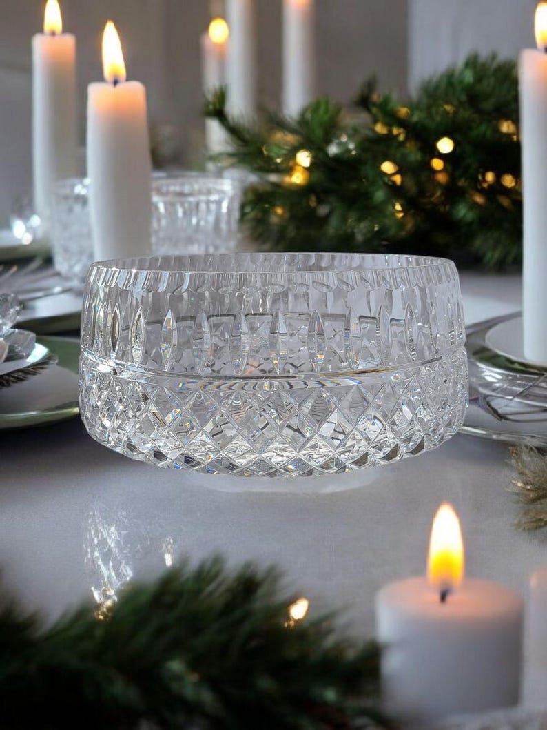 Puede incluir: Un cuenco de cristal tallado transparente con un patr&oacute;n de diamantes. El cuenco est&aacute; sobre una superficie blanca, rodeado de velas blancas encendidas y ramitas de hoja perenne, lo que sugiere un tema festivo o navide&ntilde;o. El intrincado dise&ntilde;o del cuenco capta la luz.