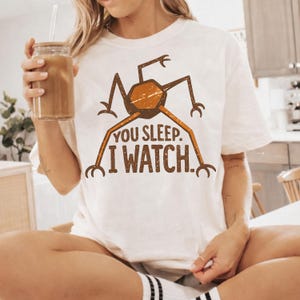 May include: White t-shirt featuring a brown and orange insect-like graphic with the text "YOU SLEEP. I WATCH." The design has a vintage, distressed look. The shirt is a casual, unisex style.