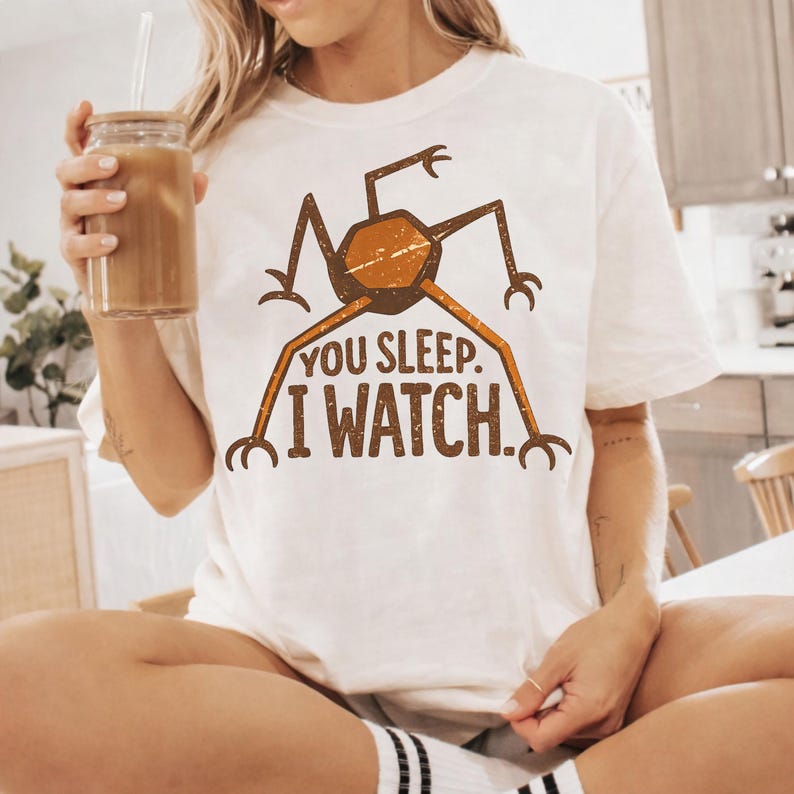 May include: White t-shirt featuring a brown and orange insect-like graphic with the text "YOU SLEEP. I WATCH." The design has a vintage, distressed look. The shirt is a casual, unisex style.