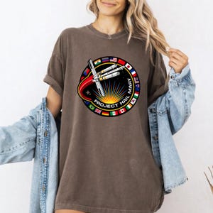 May include: Brown t-shirt featuring a circular graphic with a rocket design, the text "PROJECT HAIL MARY," and various international flags. The design includes a red arc and a sunburst pattern.