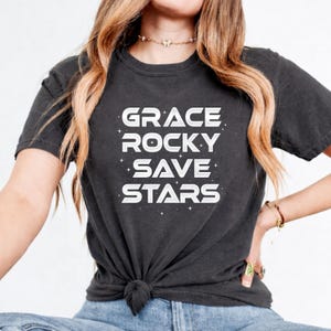 May include: Dark gray t-shirt with the words "GRACE ROCKY SAVE STARS" in white block letters. The text is arranged vertically, with small star accents. The shirt is knotted at the waist.