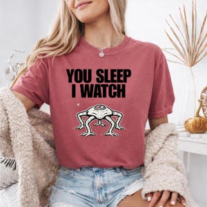 May include: A maroon t-shirt with the text "YOU SLEEP I WATCH" in black. Below the text is a white and black illustration of a creature with four legs and a single eye. The shirt is short-sleeved.