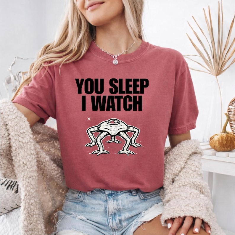 May include: A maroon t-shirt with the text "YOU SLEEP I WATCH" in black. Below the text is a white and black illustration of a creature with four legs and a single eye. The shirt is short-sleeved.