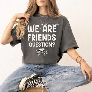 May include: Dark gray t-shirt with the text "WE ARE FRIENDS QUESTION?" in white, above a small alien illustration. The shirt also has the text "-PROJECT HAIL MARY" above the main text.