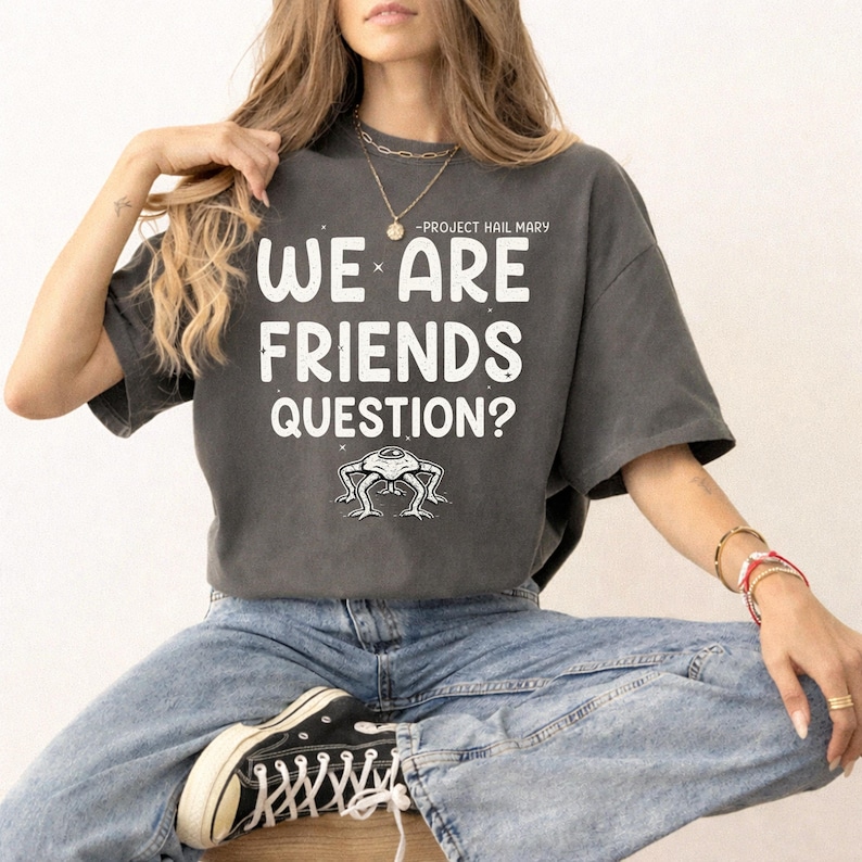 May include: Dark gray t-shirt with the text "WE ARE FRIENDS QUESTION?" in white, above a small alien illustration. The shirt also has the text "-PROJECT HAIL MARY" above the main text.