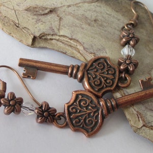 May include: A pair of copper key earrings with ornate details and clear crystal beads. The keys have a vintage style and are attached to a hook-style earring.