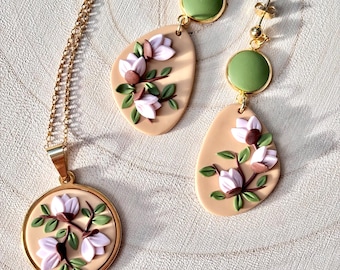 Puede incluir: Un collar y pendientes de oro con un delicado diseño floral. El colgante y los pendientes presentan una rama con flores rosas y hojas verdes sobre un fondo beige. Los pendientes también tienen un acento circular verde.