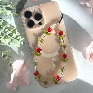May include: A clear phone case with a beaded charm attached. The charm is made of red and green glass beads in the shape of flowers. The phone case is on a white surface.