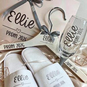 May include: A gift set featuring a light pink paper bag, a white wooden hanger, a clear glass flute, and a pair of white slippers. Each item is personalised with the name Ellie, the text PROM 2026, and a heart icon in silver or black ink.