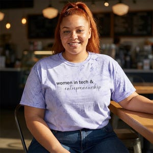 May include: A light purple tie-dye t-shirt with the text "women in tech & entrepreneurship" in black script. The shirt is worn with blue jeans. The person is sitting at a wooden table.