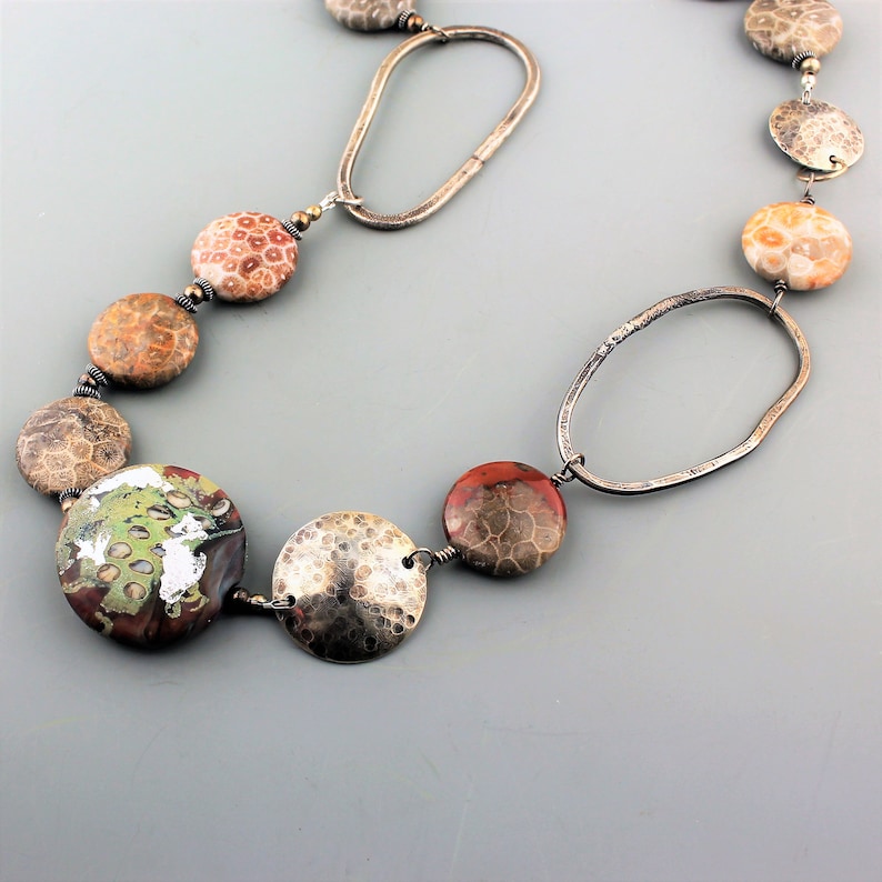 May include: A handcrafted necklace featuring large, round, fossilized stone beads in shades of brown, orange, and green. The necklace is accented with hammered silver-tone metal discs and oval links, creating a unique and earthy design.