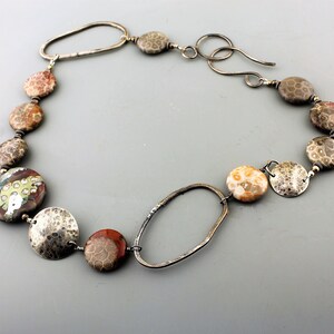 May include: A handcrafted necklace featuring a variety of natural stone beads in shades of brown, red, and cream, interspersed with hammered silver-tone metal elements. The necklace has a unique oval clasp and is designed to be worn around the neck.