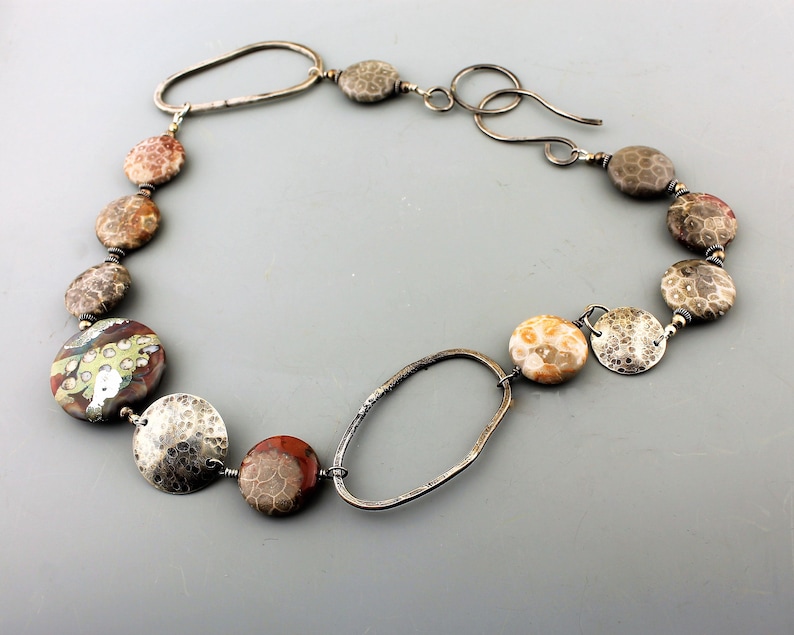 May include: A handcrafted necklace featuring a variety of natural stone beads in shades of brown, red, and cream, interspersed with hammered silver-tone metal elements. The necklace has a unique oval clasp and is designed to be worn around the neck.