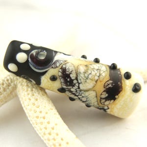 May include: A cylindrical glass bead featuring a black base with white polka dots and a central section displaying a cream-colored marbled pattern with dark brown veining. Small black raised glass dots are applied along the surface.