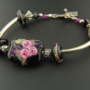 May include: A silver bangle bracelet with black and pink glass beads. The center bead features a floral design with pink roses.